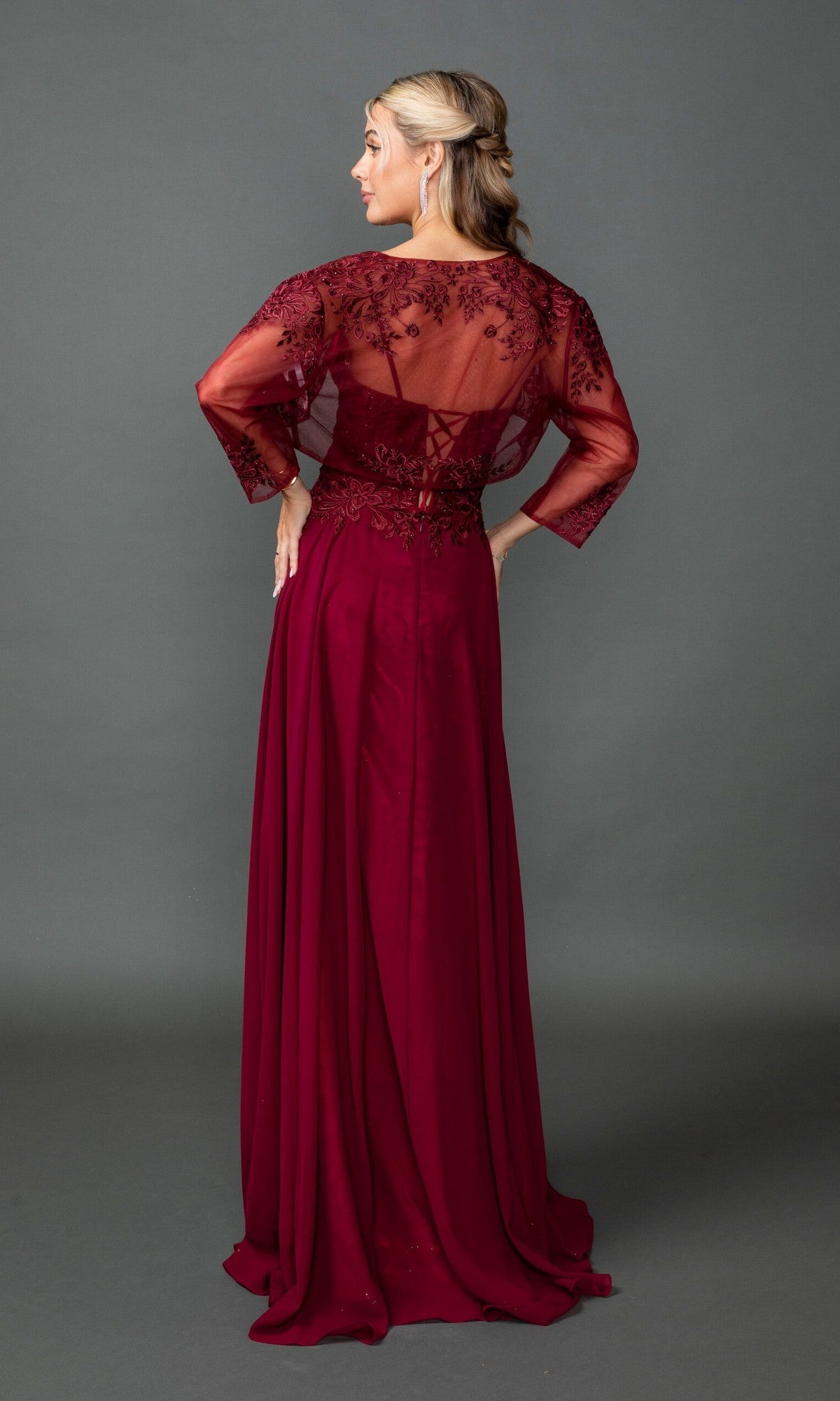 Woman wearing the Dancing Queen 4542 long prom dress in burgundy with lace embroidery on sheer sleeves and back, stands facing away against a plain gray background.
